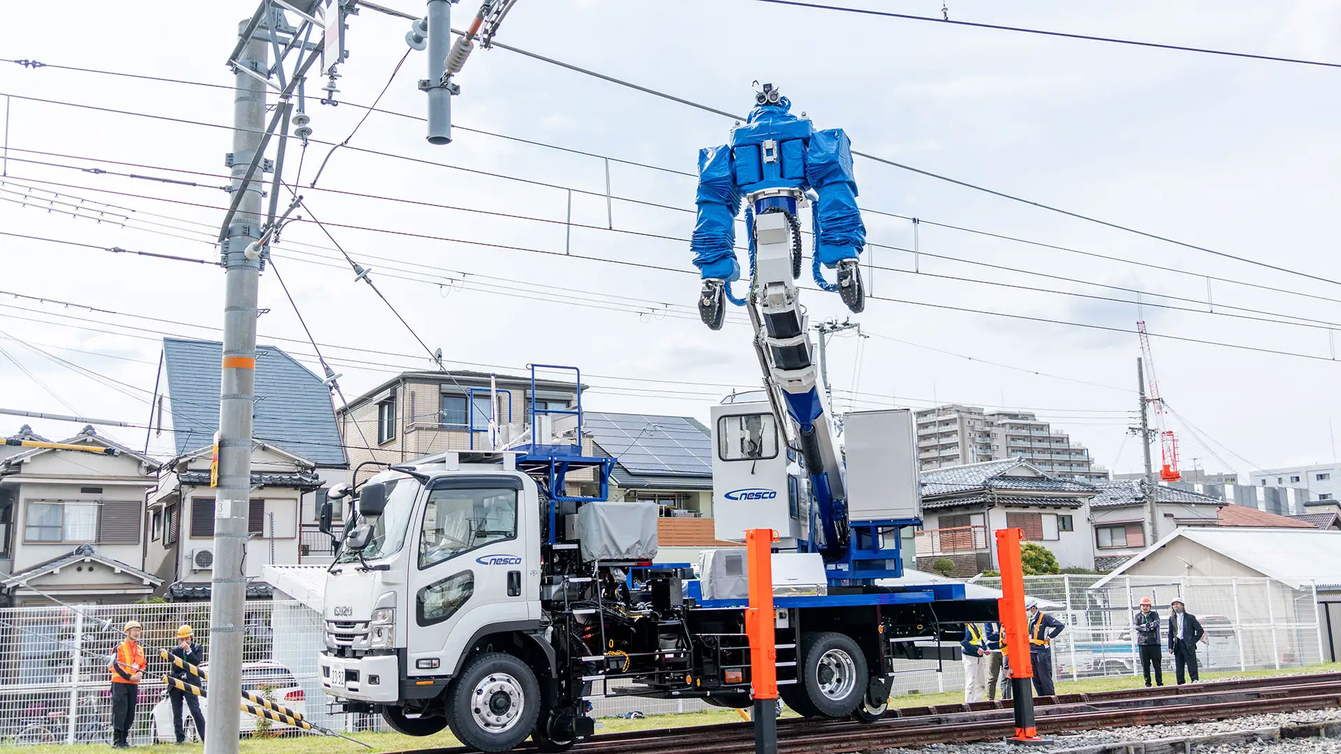 写真）多機能鉄道重機が作業している様子。人型重機は鉄道工事用車両の荷台から伸びているブーム（クレーン）に支えられており、荷台にある操縦室から遠隔操作される。